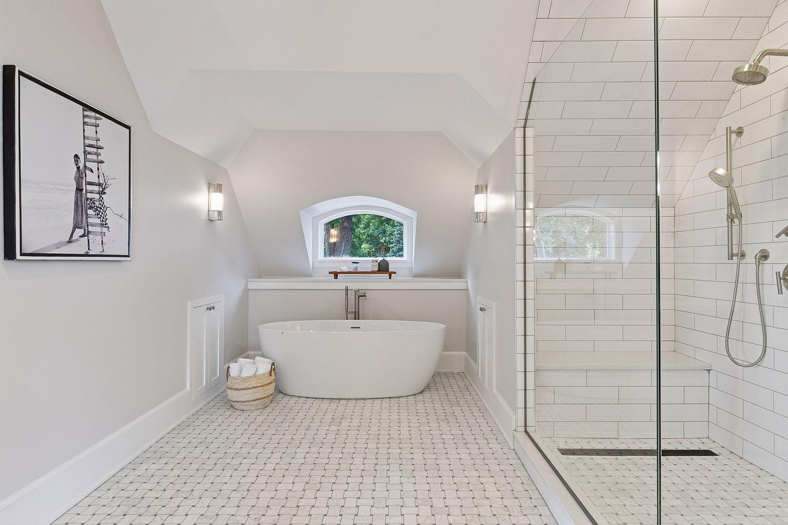 This is a bright and airy primary bathroom featuring a freestanding white bathtub positioned beneath an arched window, complemented by a glass-enclosed shower with white subway tiles. The floor is covered in a mosaic tile pattern, and the walls are painted in a neutral tone, creating a serene and spa-like atmosphere. A framed black and white print hangs on the wall, adding a touch of sophistication.
