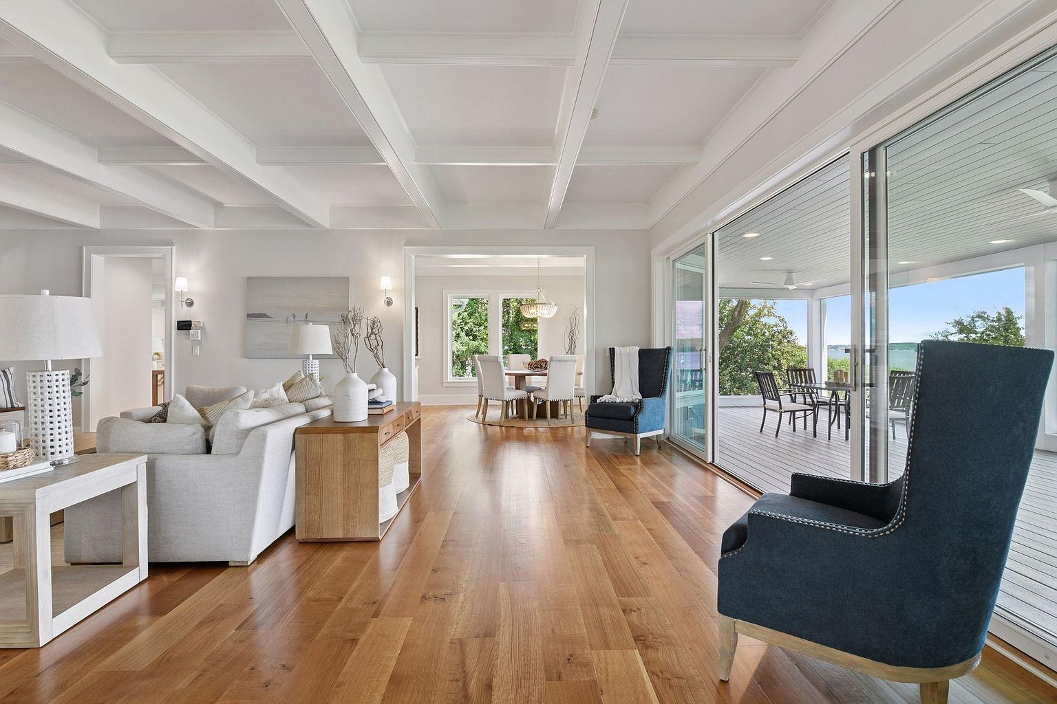 This is an interior shot of a bright and airy living room that seamlessly connects to an outdoor deck through large sliding glass doors. The room features hardwood floors, a coffered ceiling, and comfortable seating, including a light-colored sofa and two blue armchairs. The open layout flows into a dining area, creating a spacious and inviting atmosphere.