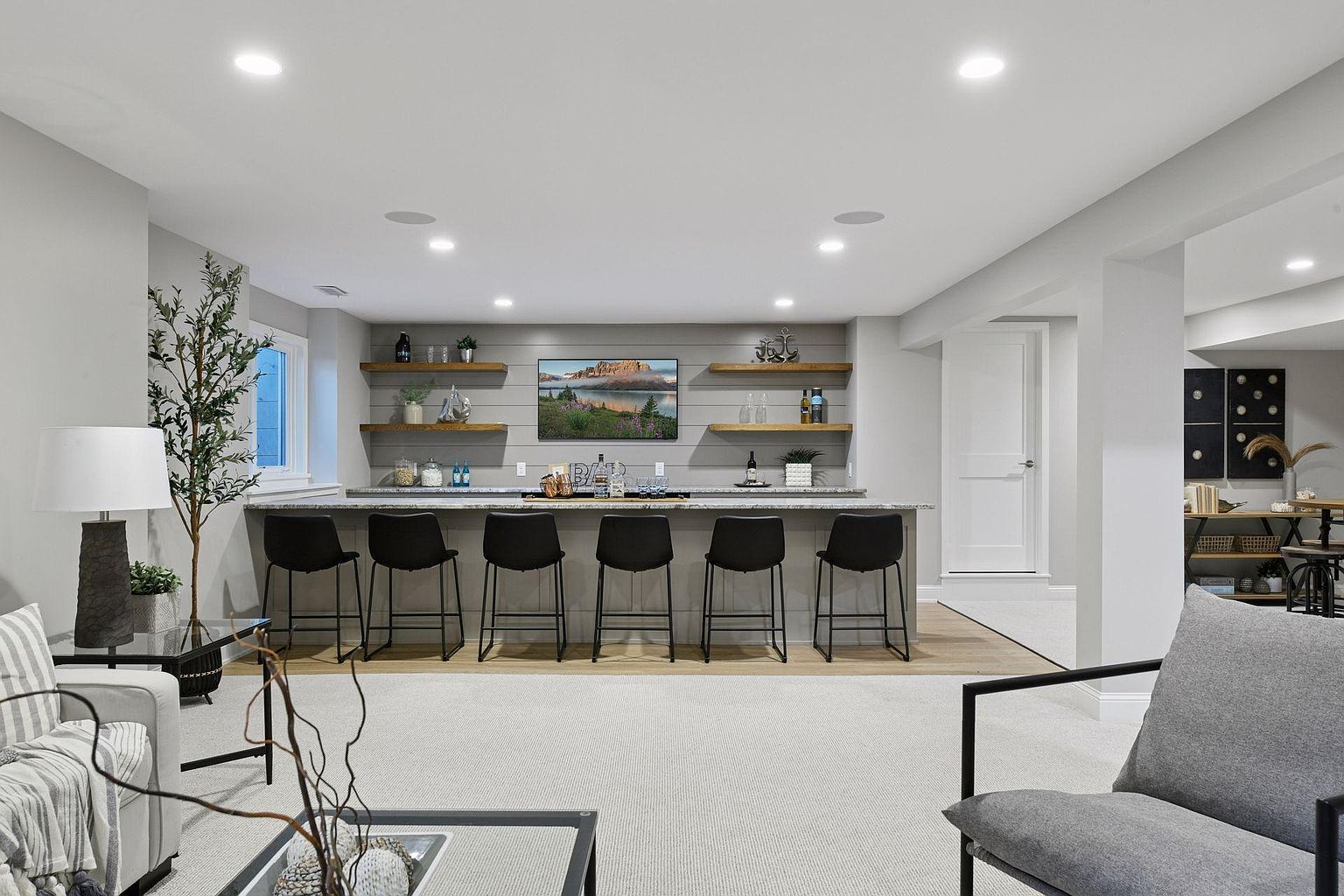 This is a well-lit basement featuring a stylish bar area with black bar stools and open shelving. A large television is mounted above the bar, and the space includes comfortable seating with a modern aesthetic. The overall impression is a finished and inviting entertainment space.
