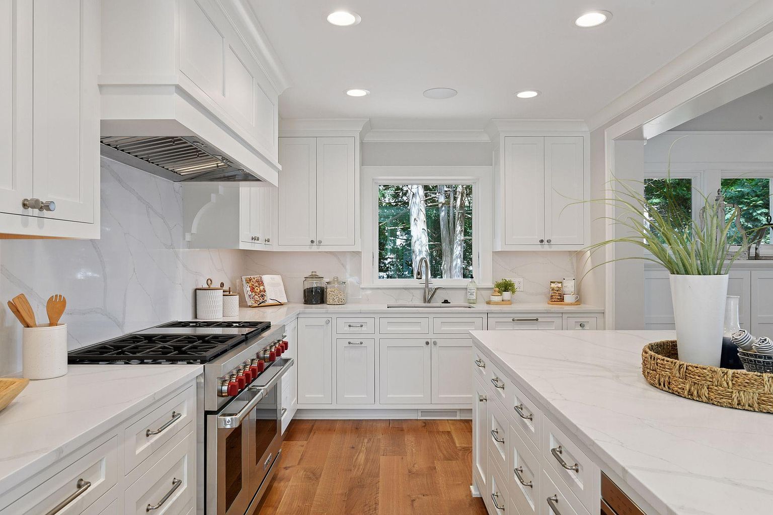 This is a bright, modern kitchen featuring white cabinetry, stainless steel appliances, and light countertops. The kitchen has a large island and a window above the sink that provides natural light. The hardwood floors add warmth to the space, creating a clean and inviting atmosphere.