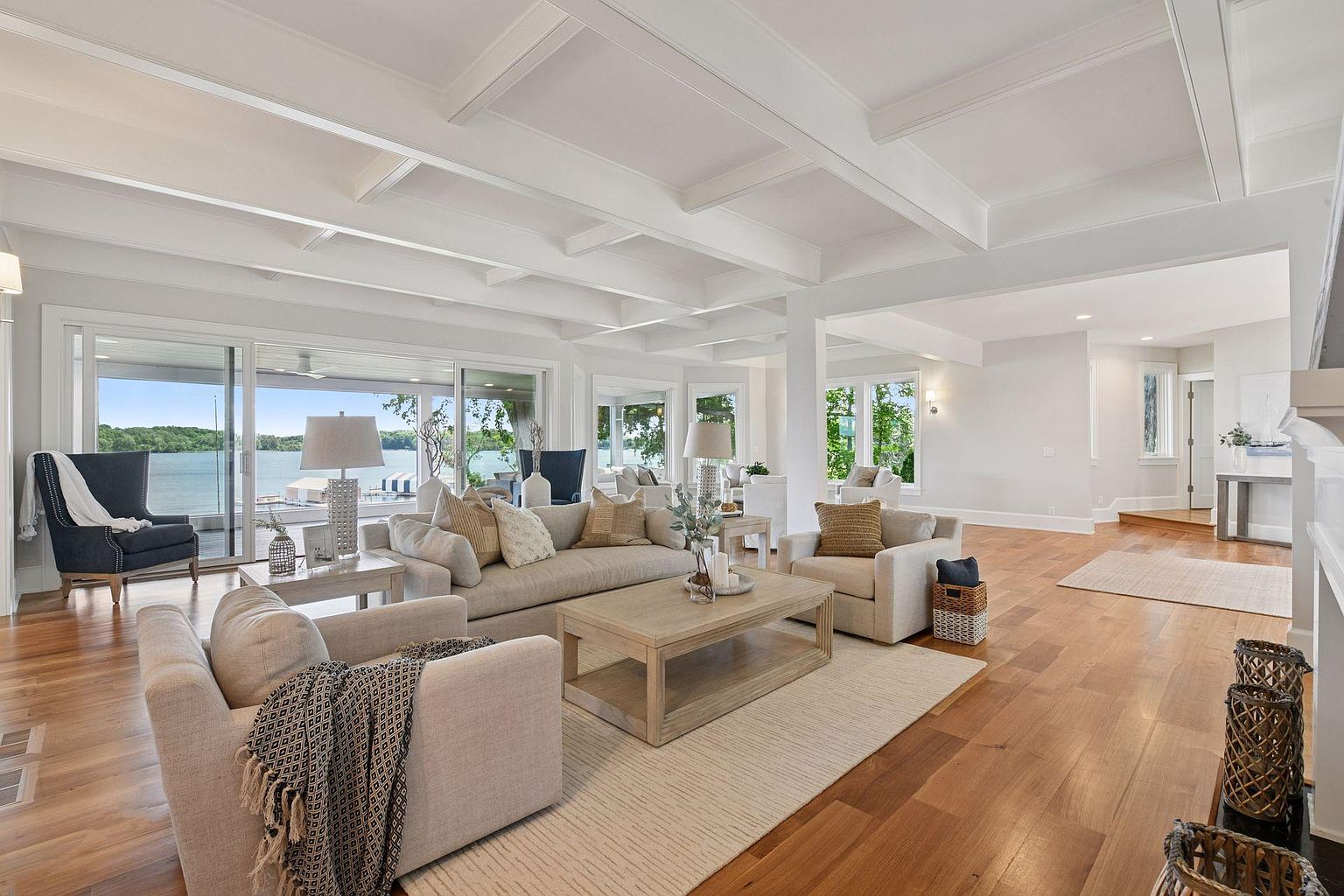 This is a bright and airy living room with a view of the lake. The room features hardwood floors, white coffered ceilings, and neutral-toned furniture, including a large sofa and armchairs. Large windows and sliding glass doors provide ample natural light and access to the outdoors, creating a seamless indoor-outdoor living experience.