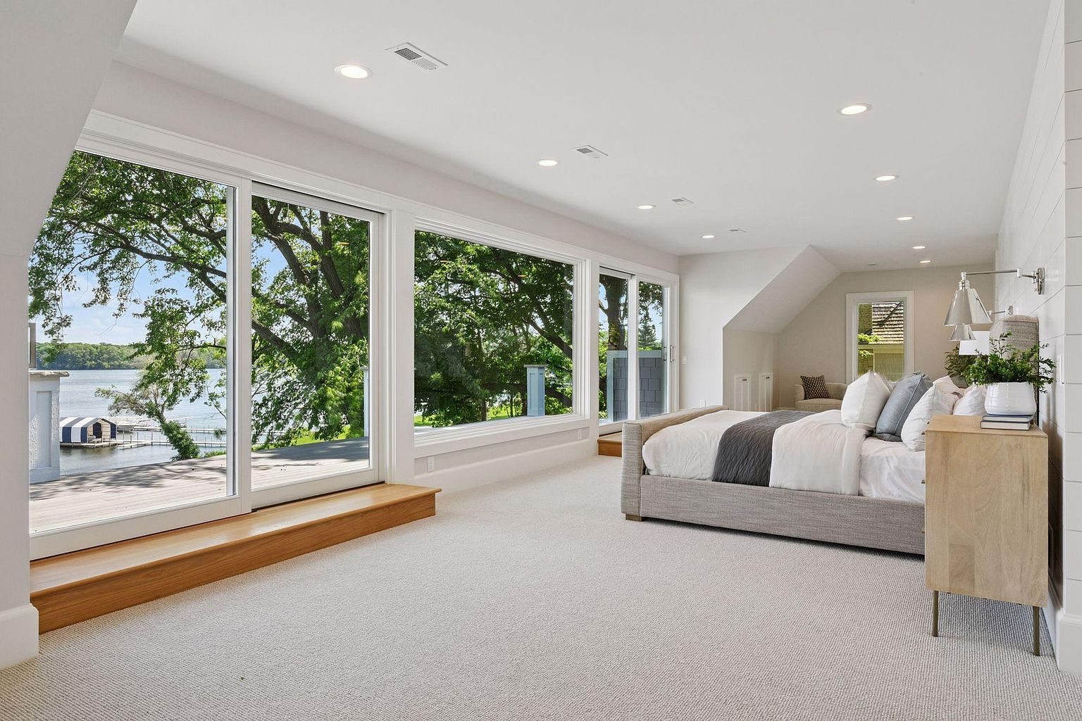 This is a bright and spacious primary bedroom with a wall of windows offering a scenic view of the water and lush greenery. The room features neutral tones, light-colored carpet, and a minimalist design, creating a serene and inviting atmosphere. A platform bed with a gray frame and a light wood nightstand add to the room's modern aesthetic.