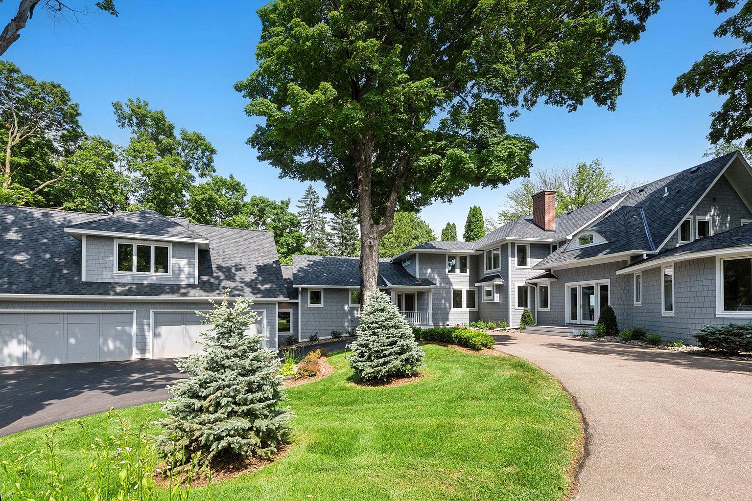 This is a front view of a large, luxurious home with gray siding and a dark gray roof. The property features a well-manicured lawn, mature trees, and a circular driveway leading to the house and a detached garage. The overall impression is one of elegance and privacy.