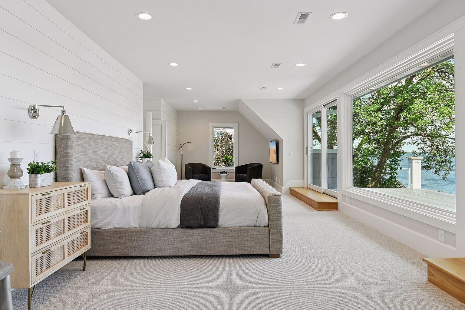 This is a bright and airy primary bedroom featuring a neutral color palette and modern design. The room includes a large bed with a textured headboard, a stylish nightstand, and a sitting area with armchairs. Large windows offer a view of the outdoors, enhancing the serene and luxurious atmosphere.
