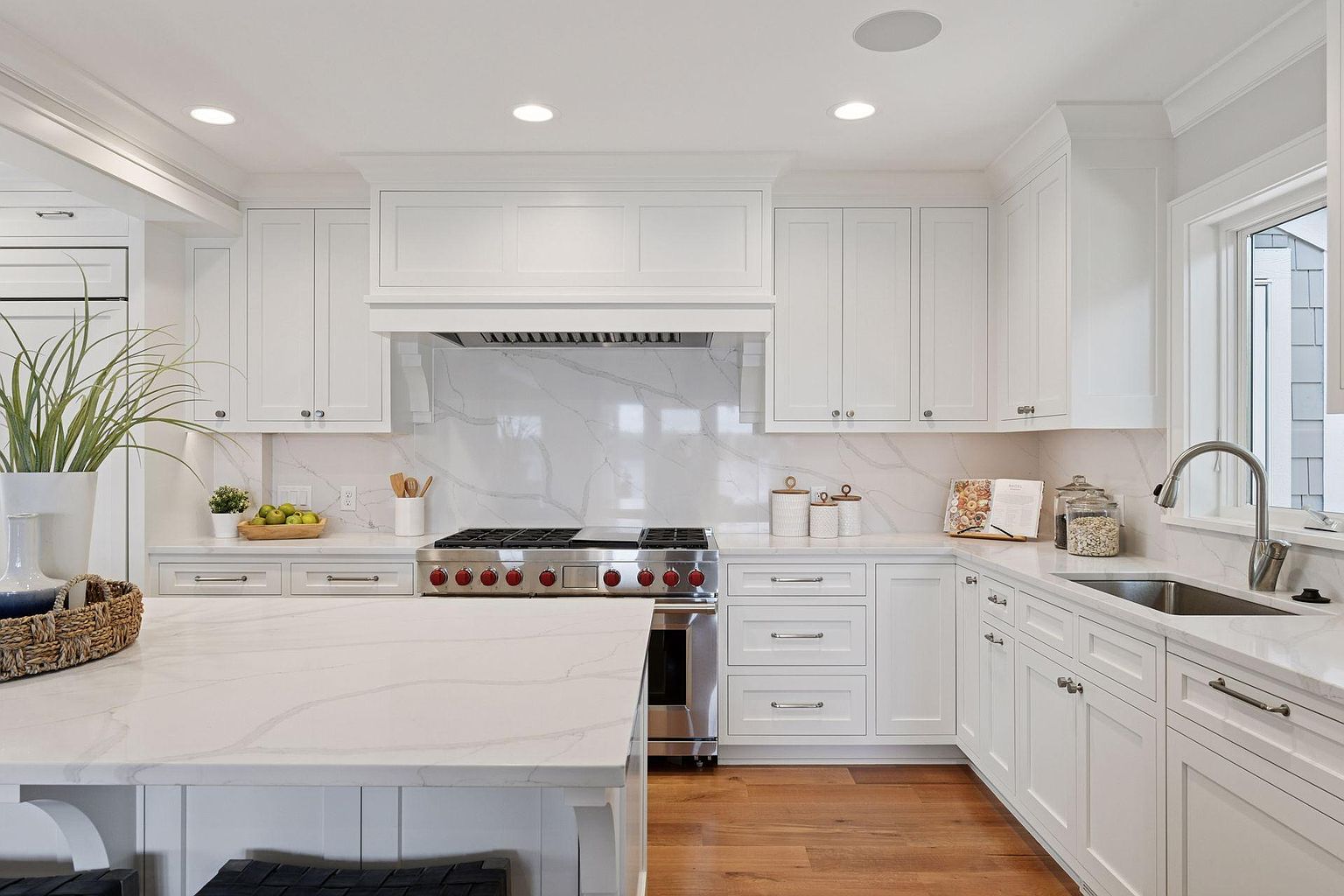 This is a bright and modern kitchen featuring white cabinetry, stainless steel appliances, and a marble countertop island. The kitchen is well-lit with recessed lighting and natural light from a window above the sink. The hardwood floors add warmth to the space, creating an inviting atmosphere.