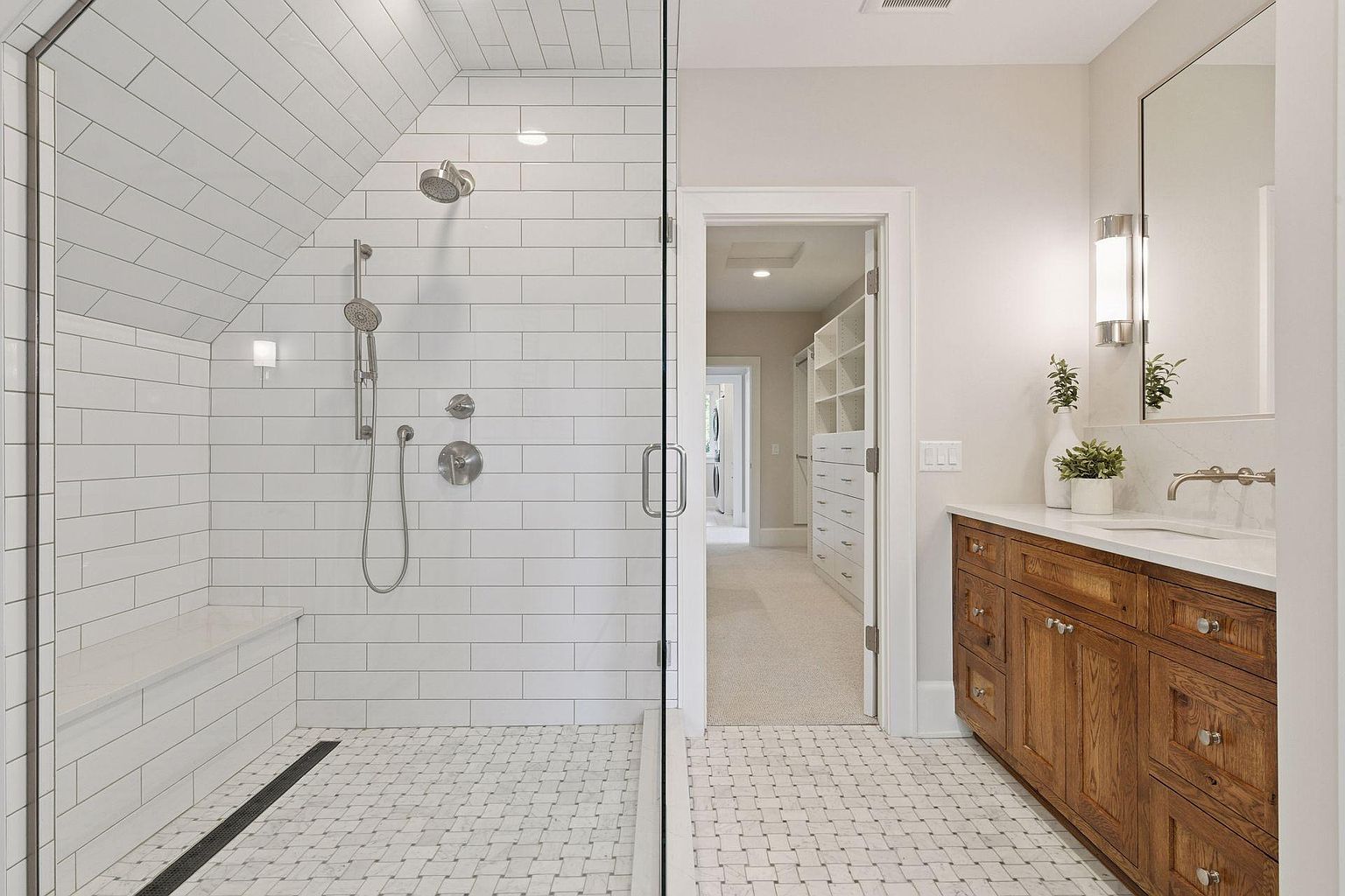 This is a well-lit primary bathroom featuring a walk-in shower with white subway tile and a built-in bench. The floor is covered in a mosaic tile pattern, and a wooden vanity with a white countertop and modern fixtures is visible. An open doorway leads to a walk-in closet, creating a sense of spaciousness and luxury.