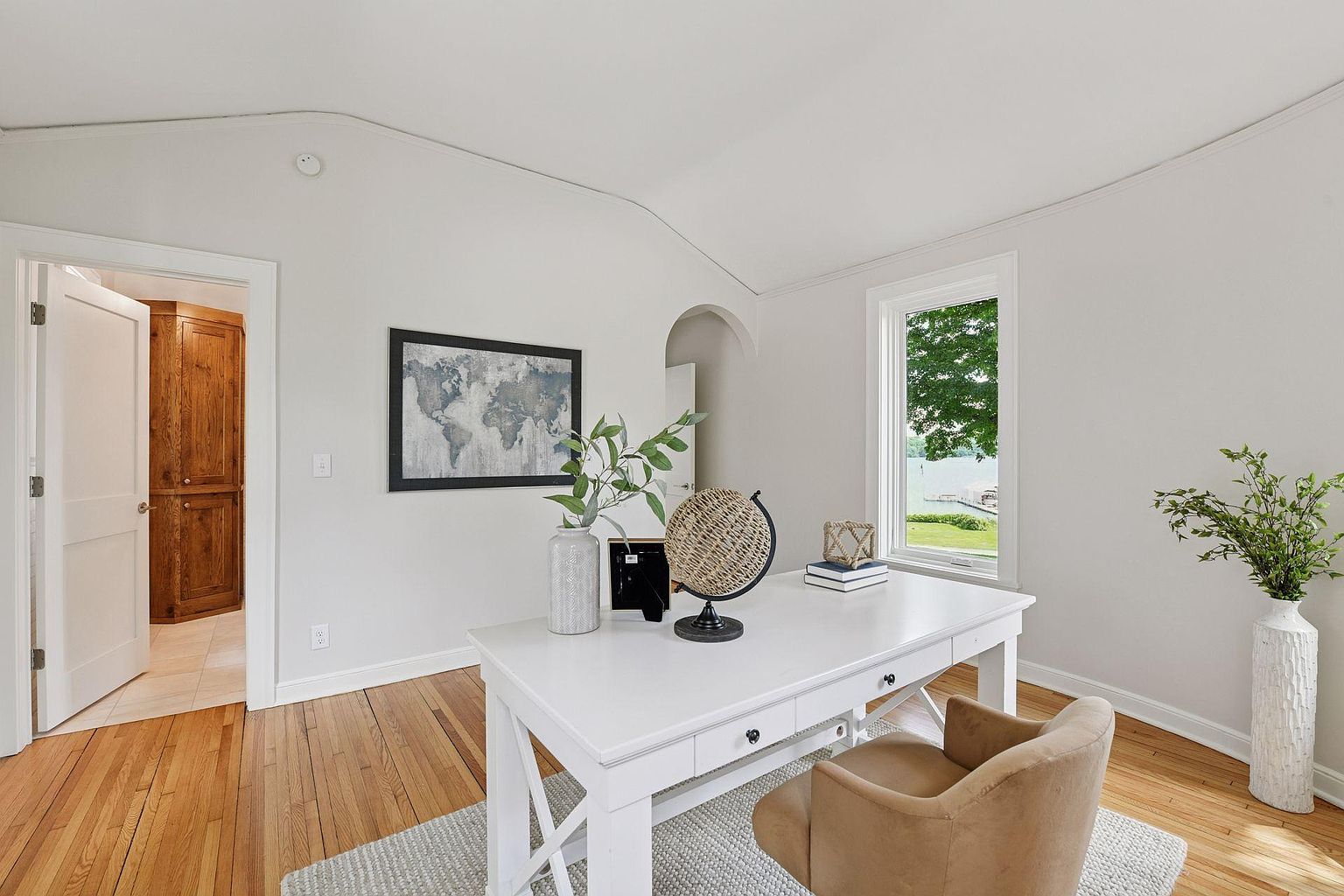 This is an interior shot of a bright and airy home office. The room features a white desk with decorative items, a comfortable chair, and a large window offering a view of the outdoors. A world map hangs on the wall, and the hardwood floors add warmth to the space, creating an inviting and productive atmosphere.