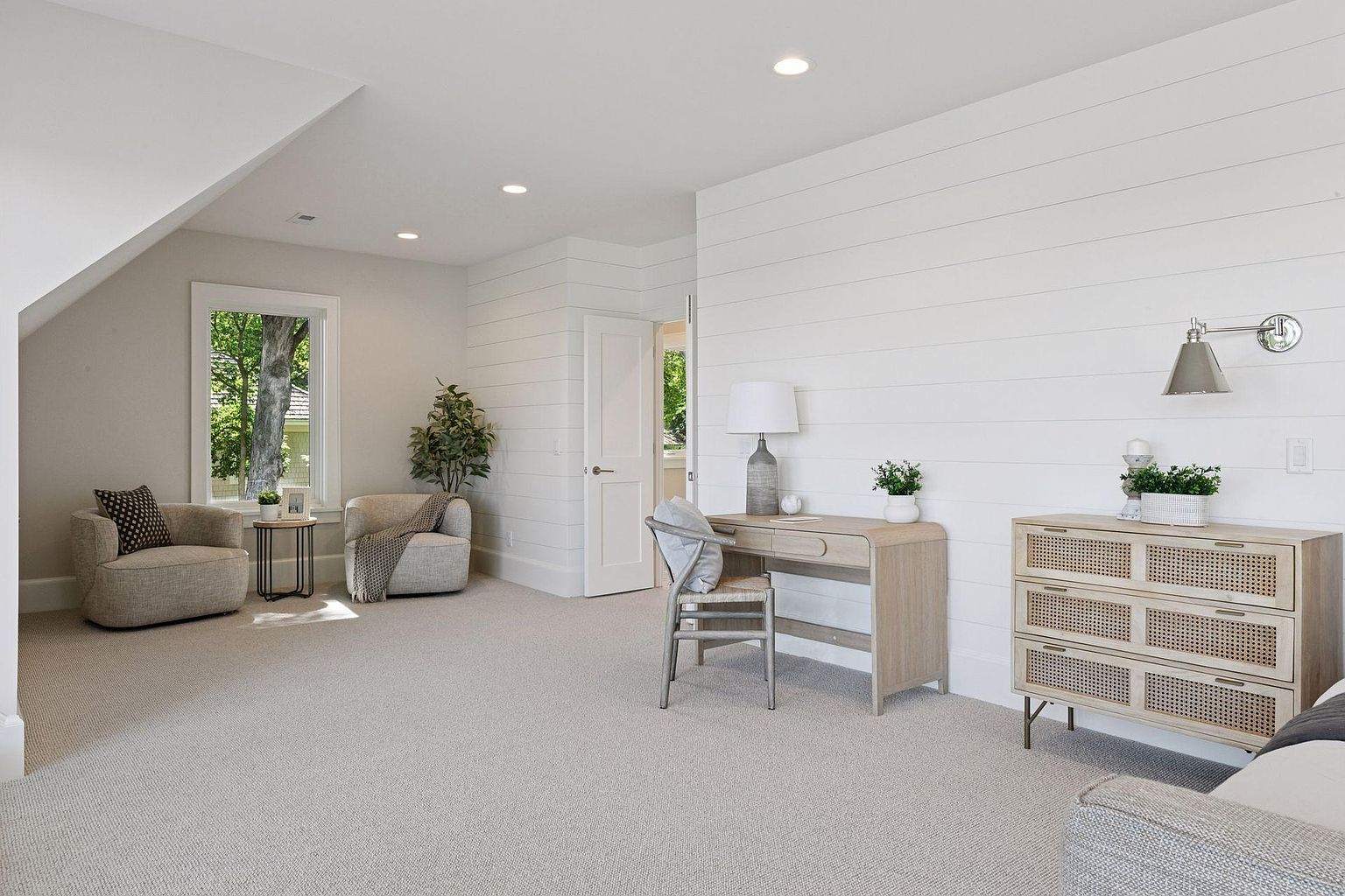 This is a bright and airy guest bedroom featuring a neutral color palette and shiplap accent walls. The room includes two comfortable armchairs by a window, a desk with a chair, and a dresser with a modern wall sconce. The overall impression is clean, cozy, and inviting, perfect for a relaxing retreat.