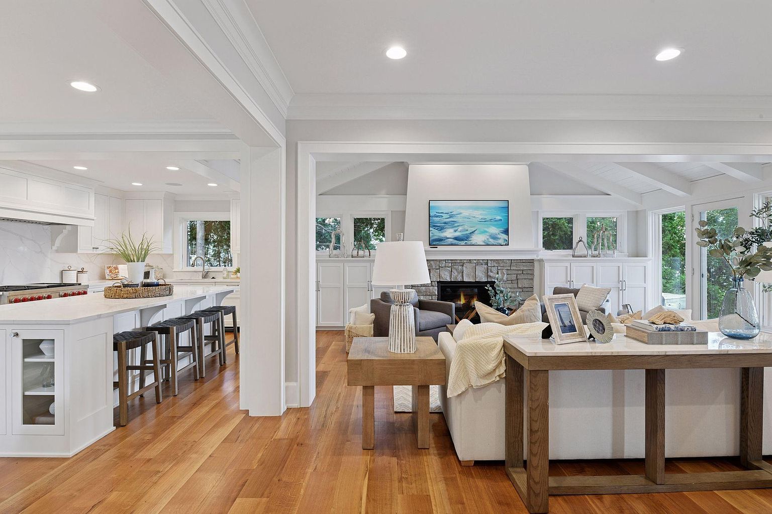 This is an interior shot showcasing an open-concept living room and kitchen area. The living room features a stone fireplace with a mounted television, comfortable seating, and stylish decor. The kitchen includes a large island with bar stools, white cabinetry, and modern appliances, creating a seamless and inviting living space.