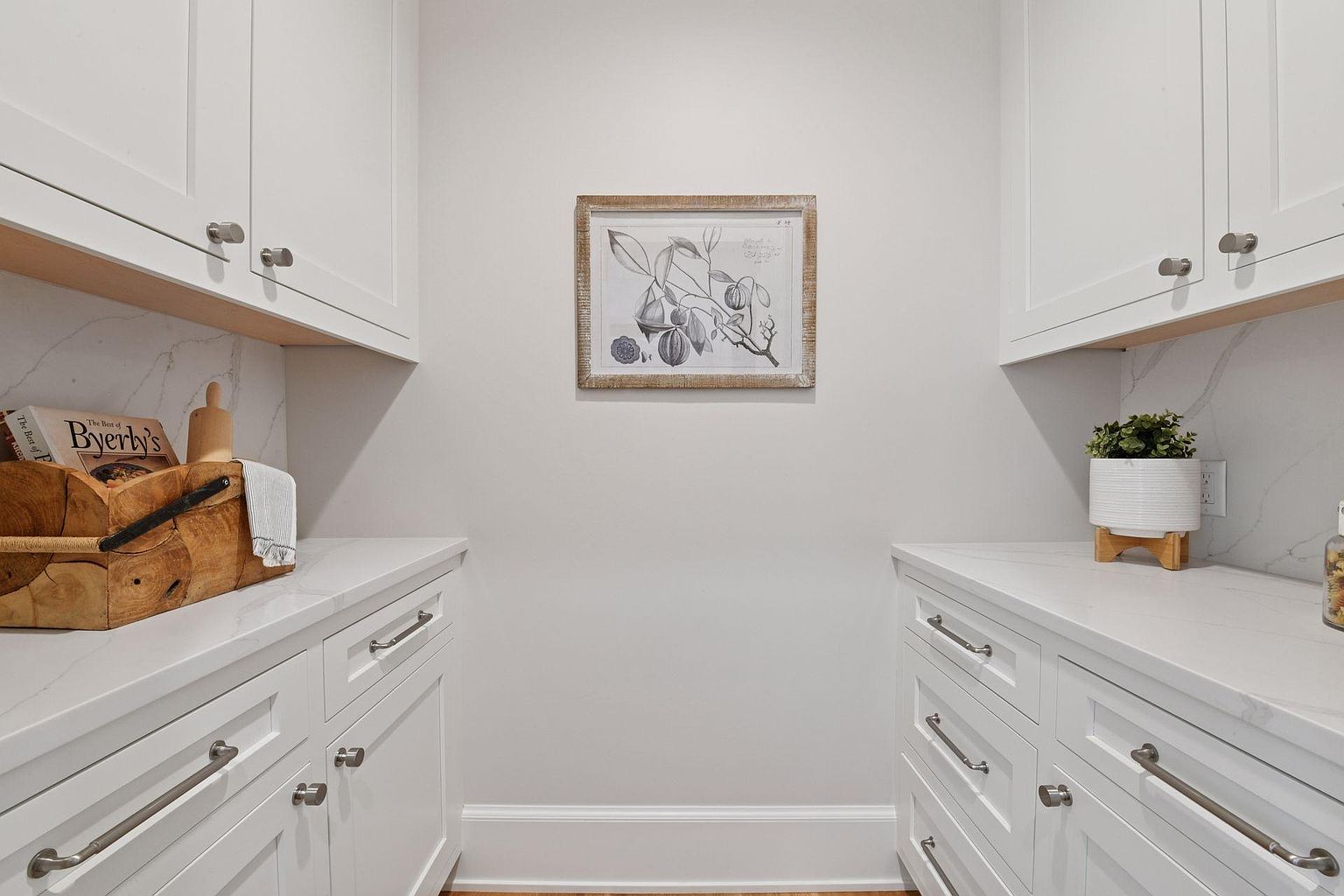 This is an interior shot of a well-organized pantry. White cabinets and drawers line both sides of the space, topped with white countertops that have a subtle marble pattern. A framed botanical print hangs on the wall, adding a touch of elegance to the functional space.