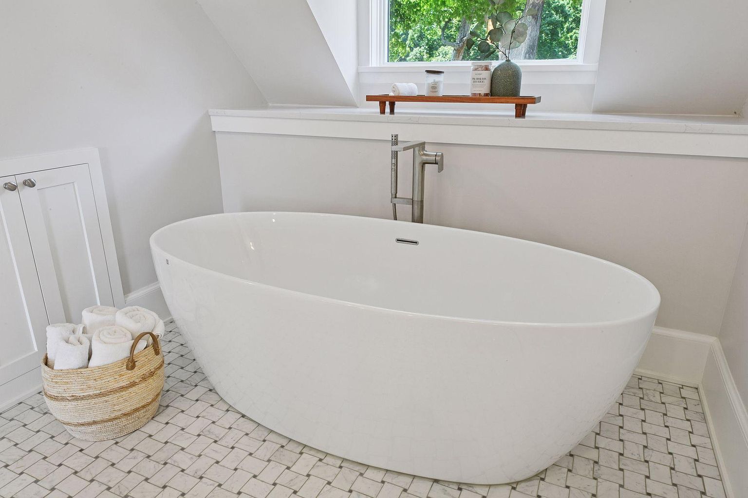 This is a bright and airy primary bathroom featuring a freestanding white bathtub as the focal point. The room is decorated in neutral tones, with white walls and light-colored tile flooring. A window provides natural light, and a wooden shelf above the tub holds decorative items, creating a spa-like atmosphere.