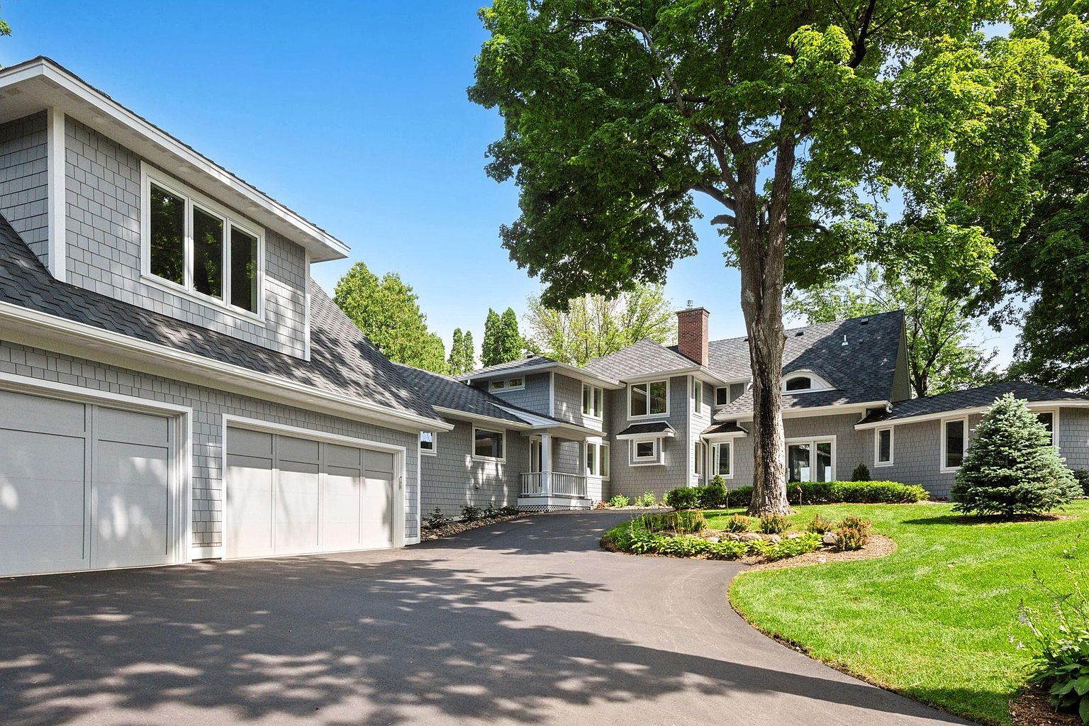 This is a front exterior view of a large, well-maintained home with gray shingle siding and a dark roof. The property features a spacious driveway leading to a multi-car garage, mature trees providing shade, and a manicured lawn. The architectural style suggests a blend of traditional and contemporary elements, creating an inviting and upscale impression.