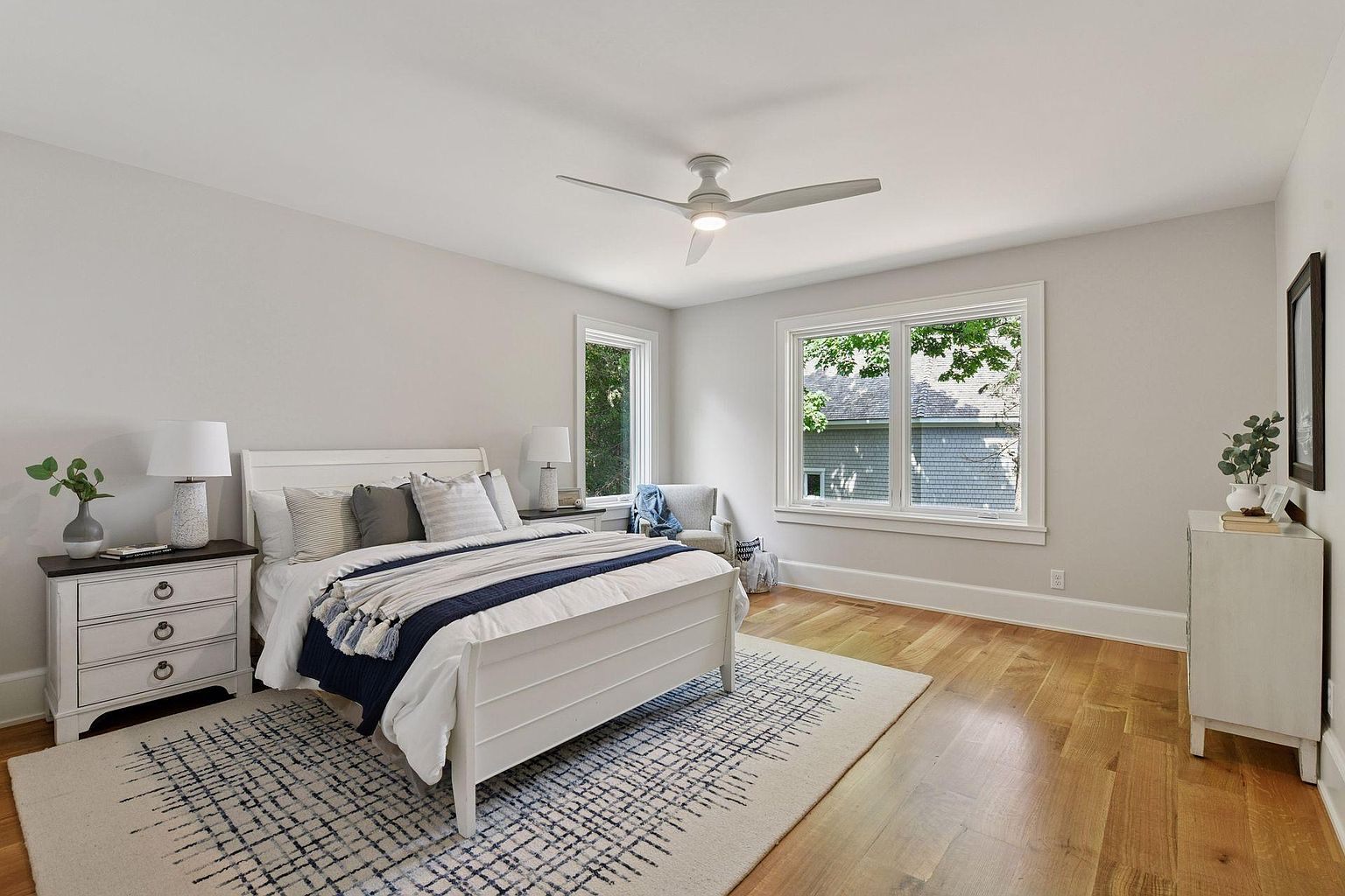 This is a bright and airy primary bedroom featuring a white bed with a blue and white throw blanket, complemented by a neutral-toned rug. The room has hardwood floors and is illuminated by natural light streaming through two windows. A ceiling fan adds to the comfort of the space.