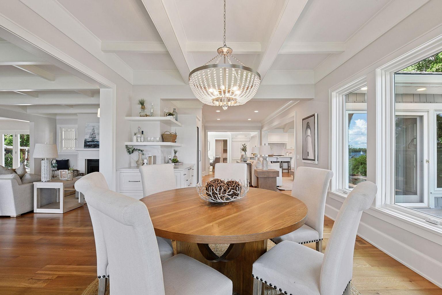 This is an interior shot of a dining room featuring a round wooden table surrounded by four upholstered chairs. A crystal chandelier hangs above the table, and natural light floods the room through a large window. The room is decorated in a neutral color palette, creating a bright and inviting atmosphere.