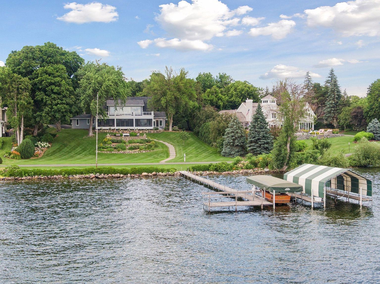 This aerial view showcases a stunning lakeside property featuring multiple homes nestled among lush greenery. A private dock extends into the water, complete with boat storage. The manicured lawns and gardens add to the estate's appeal, creating a serene and luxurious atmosphere.