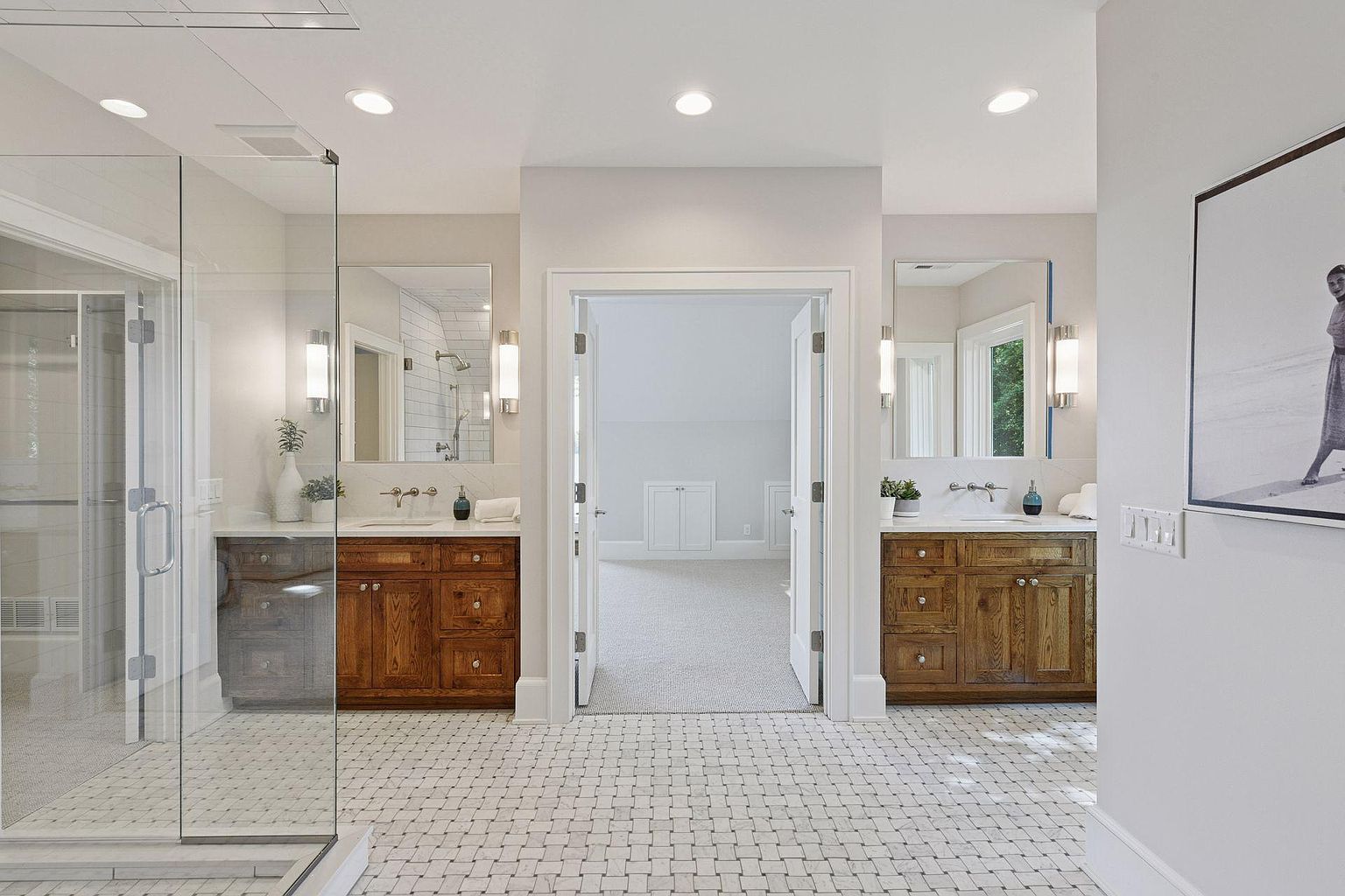 This is a well-lit primary bathroom featuring dual wooden vanities with marble countertops and modern sconces. The floor is tiled in a basketweave pattern, and a glass-enclosed shower is visible to the left. An open doorway leads to another room, creating a sense of spaciousness and luxury.