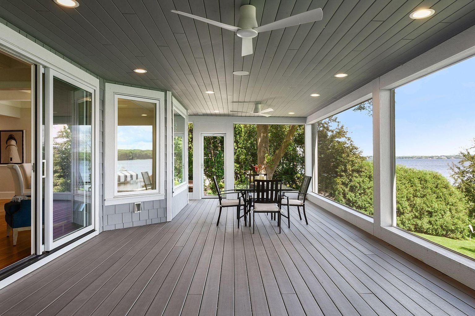 This image showcases a spacious outdoor patio or deck area, featuring a dining set with chairs and a table, suggesting an ideal space for outdoor meals and gatherings. The area is enclosed with large windows offering views of the surrounding landscape, including trees and a body of water, creating a serene and inviting atmosphere. The ceiling is finished with wood paneling and recessed lighting, adding a touch of elegance.
