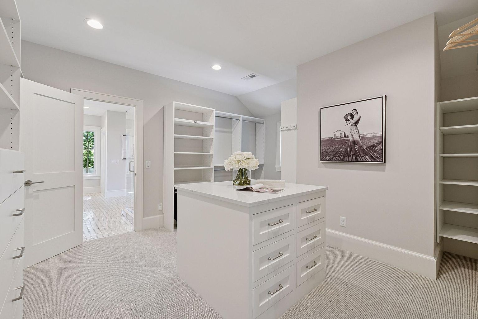 This is a well-organized walk-in closet featuring custom shelving and drawer units. A central island provides additional storage and countertop space, topped with a vase of white flowers and a book. The room is brightly lit with recessed lighting and has a neutral color palette, creating a clean and luxurious feel.