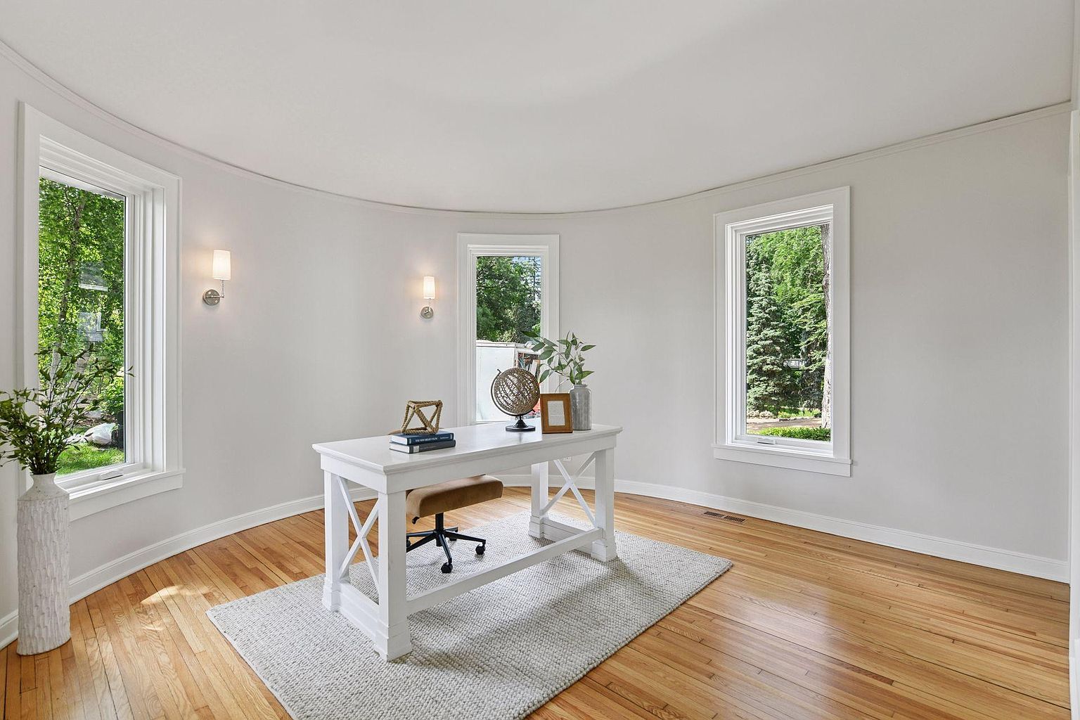 This is an interior shot of a bright and airy home office. The room features a curved wall with three large windows offering views of the outdoors, a white desk with a chair, and a light-colored rug on hardwood floors. The overall impression is clean, modern, and conducive to productivity.