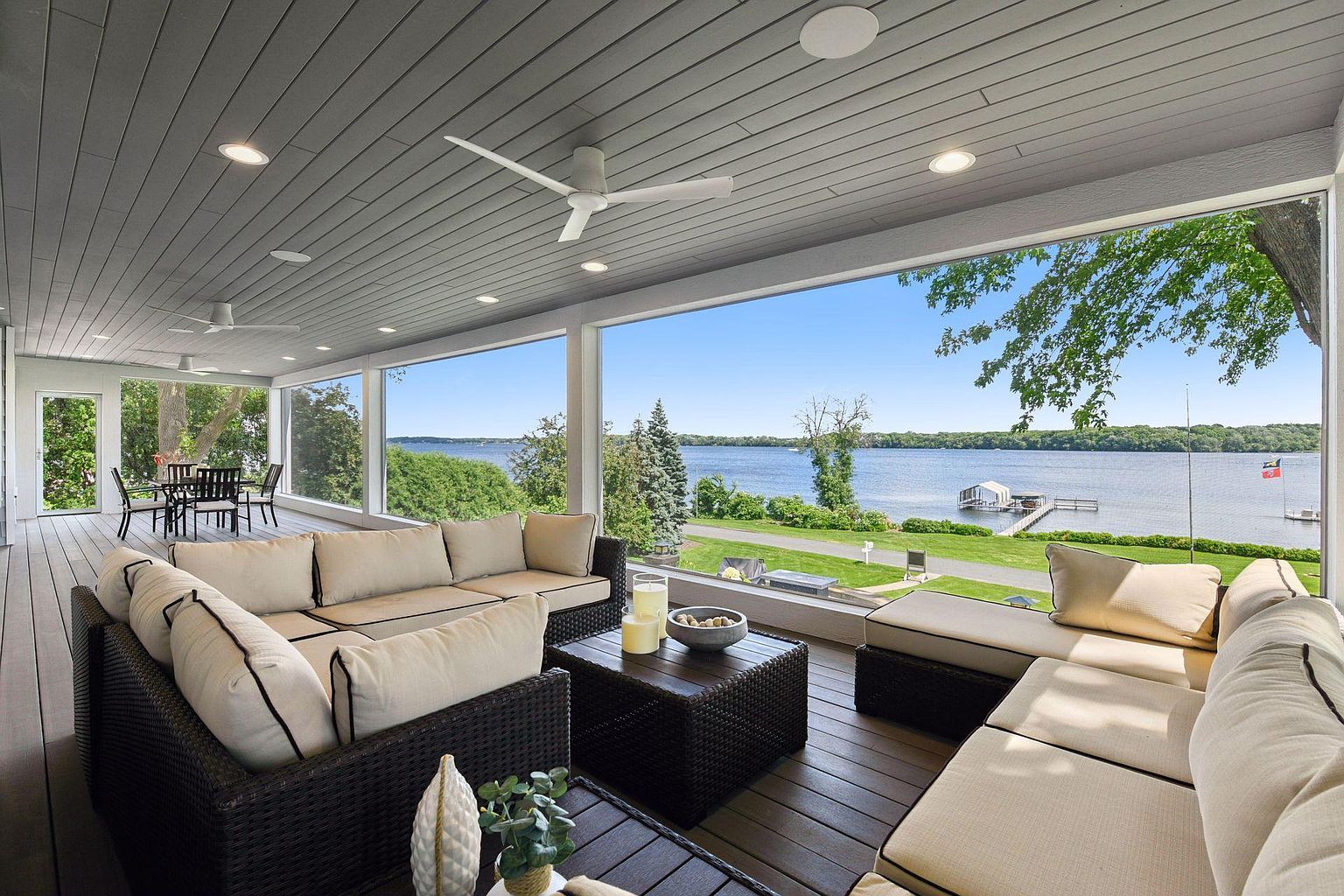 This is a luxurious outdoor patio or deck area with comfortable wicker furniture and plush cushions, offering a stunning view of a lake. The space is covered with a gray ceiling and features ceiling fans and recessed lighting, creating a relaxing and inviting atmosphere. Large windows provide an unobstructed view of the water and surrounding landscape, enhancing the property's appeal.