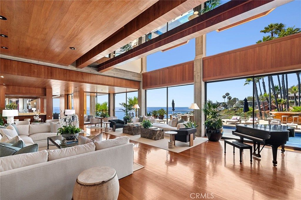 This expansive, open-concept living area features warm wood-paneled ceilings and polished hardwood floors, creating a sophisticated and airy atmosphere. The space is defined by massive, tall windows that frame panoramic ocean views and provide seamless access to the outdoor pool area. A grand piano sits prominently near the glass, while plush, neutral-toned seating arrangements are organized to take full advantage of the natural light and coastal scenery.