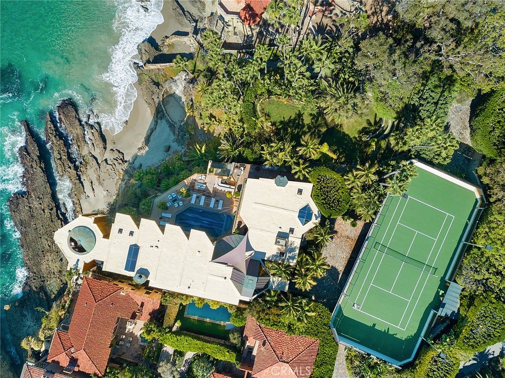This high-angle aerial view captures a sprawling coastal estate perched directly above a rocky shoreline and the ocean. The property features a large tennis court, a swimming pool nestled near the cliff edge, and multiple structures with terracotta-style roofing surrounded by lush, mature landscaping. The perspective emphasizes the dramatic integration of luxury living with the rugged natural beauty of the coastline.