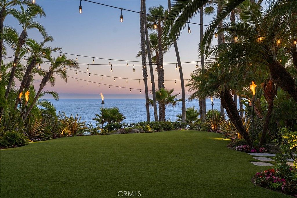 This expansive, manicured lawn overlooks a stunning ocean view at dusk, framed by lush palm trees and tropical landscaping. String lights are draped across the yard, creating an inviting atmosphere, while decorative tiki torches add a warm glow to the perimeter. The scene captures a serene and luxurious outdoor living space perfect for evening entertaining.