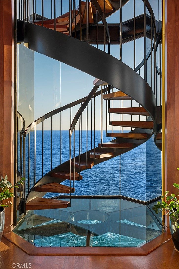 This striking interior space features a spiral staircase with wooden treads and a dark metal railing, set against a backdrop of floor-to-ceiling glass walls that offer an expansive, unobstructed view of the ocean. The floor beneath the staircase is also constructed of glass, creating a dramatic, transparent effect that emphasizes the coastal setting. The design is modern and architectural, highlighting the seamless integration between the home's interior and the surrounding seascape.