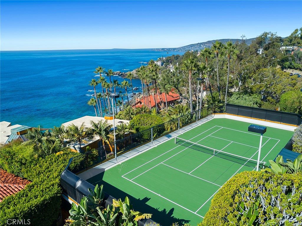 This high-angle aerial view captures a private tennis court situated on a coastal cliffside overlooking the ocean. The scene features lush greenery, palm trees, and a glimpse of a nearby residence with a terracotta-tiled roof, all set against a vast, clear blue sky and sea. The perspective emphasizes the property's exclusive location and seamless integration with the natural landscape.