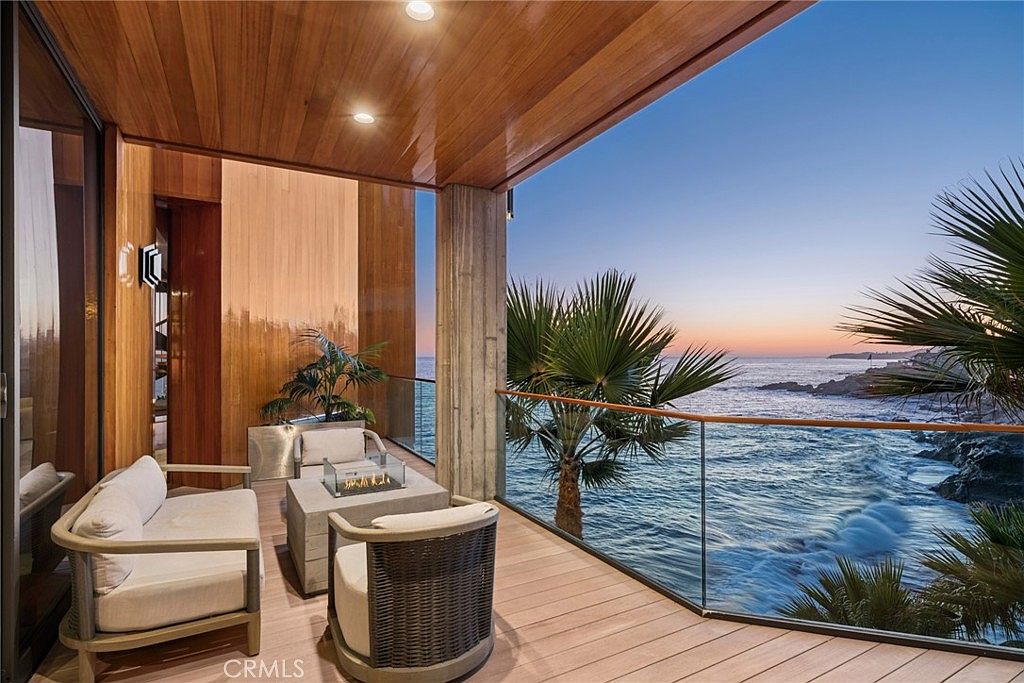 This luxurious balcony offers a seamless indoor-outdoor living experience with wood-paneled ceilings and a glass railing that provides unobstructed views of the ocean. The space is furnished with a comfortable seating area and a central fire feature, creating an inviting atmosphere for relaxation. The perspective captures the serene sunset over the water, highlighting the sophisticated design and prime coastal location.