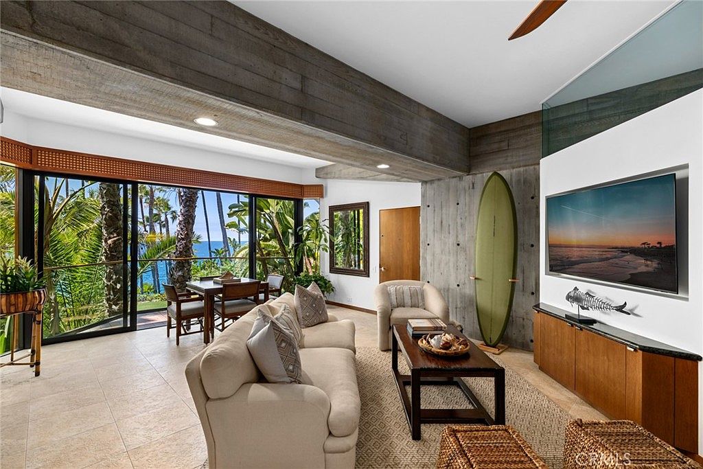 This bright living room features a coastal-inspired aesthetic with a neutral sofa, a dark wood coffee table, and a surfboard displayed against a textured concrete wall. Large sliding glass doors open to a balcony with a dining set, offering a scenic view of palm trees and the ocean beyond. The space is defined by exposed wooden ceiling beams and light-colored tile flooring, creating an airy and relaxed atmosphere.