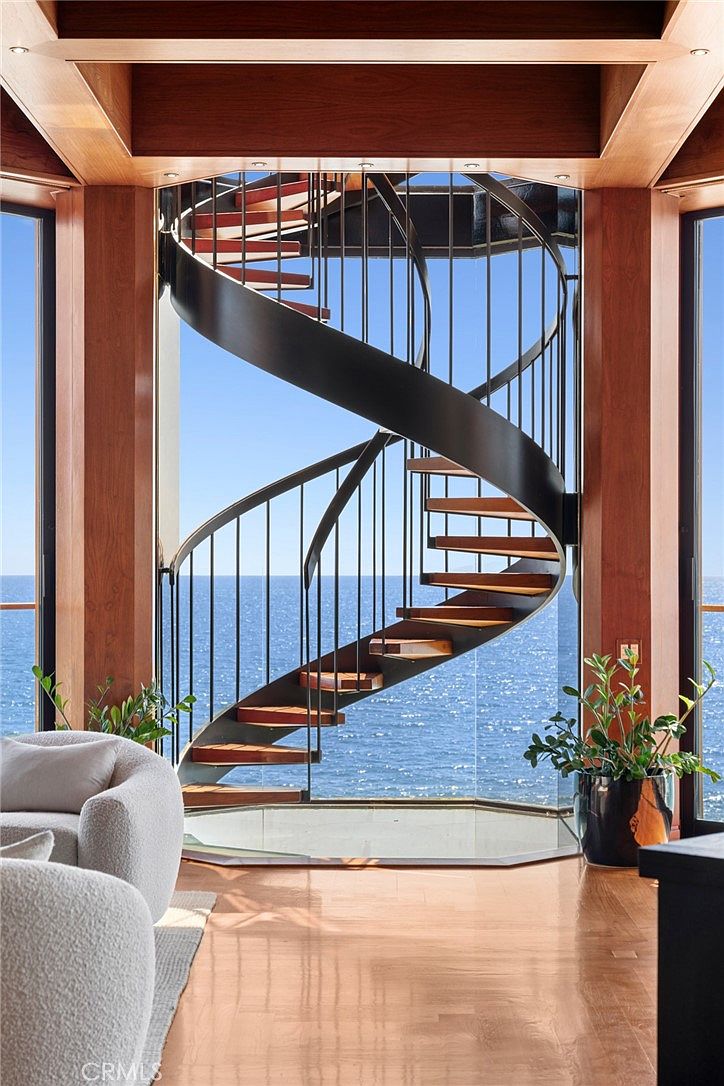 This interior space features a striking spiral staircase with wooden treads and a dark metal railing, positioned in front of expansive floor-to-ceiling glass panels that frame a panoramic ocean view. The room is framed by warm-toned wood columns and beams, creating a sophisticated and airy atmosphere. A plush, light-colored armchair and a potted plant in the foreground add a sense of comfort and scale to this dramatic architectural focal point.