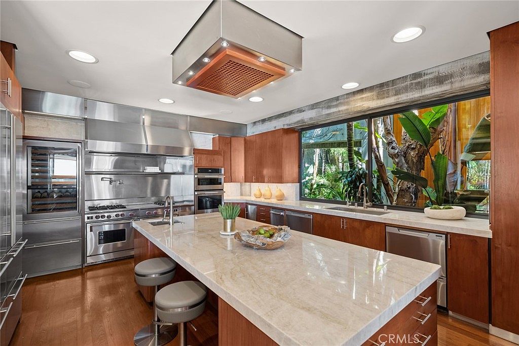 This modern kitchen features warm wood cabinetry paired with stainless steel appliances and a large central island topped with a light-toned stone surface. A prominent stainless steel range hood hangs above the island, while a wide window provides a lush, green view of the outdoors. The space is illuminated by recessed lighting, creating a bright and inviting atmosphere with a contemporary aesthetic.