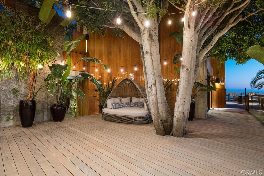 This expansive wooden deck features a large, rounded wicker daybed nestled beneath a mature tree, creating a serene and inviting outdoor lounge area. String lights are draped overhead, casting a warm glow against the vertical wood-paneled wall of the home. The space offers a seamless transition to an open-air setting with a view of the ocean in the distance, emphasizing a luxurious, resort-like atmosphere.