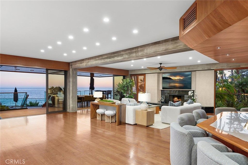 This expansive, open-concept living area features a seamless transition to an outdoor deck overlooking the ocean through large glass sliding doors. The room is anchored by a central fireplace with a stone-clad chimney breast and is furnished with plush, light-toned seating arranged around a central coffee table. A warm, wood-toned ceiling structure and polished wood flooring create a sophisticated, airy atmosphere that emphasizes the panoramic coastal views.