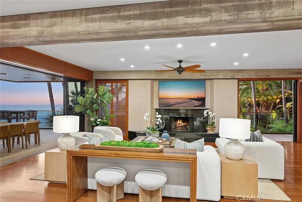This expansive living room features a warm, contemporary design with rich wood flooring and a prominent fireplace as the central focal point. The space is seamlessly connected to an outdoor deck through large sliding glass doors, offering panoramic ocean views on one side and lush greenery on the other. The room is furnished with plush, light-colored seating and a console table, creating an inviting atmosphere that blends indoor comfort with the natural beauty of the coastal setting.