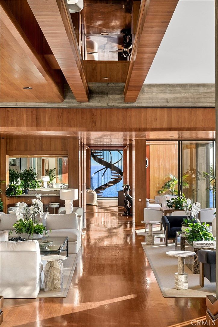 This expansive, open-concept living area features warm wood-paneled walls and ceilings, creating a sophisticated and cohesive aesthetic. The space is furnished with plush white seating and elegant side tables, leading the eye toward a striking spiral staircase that frames a stunning ocean view. The polished wood flooring reflects the natural light, enhancing the airy and luxurious atmosphere of this coastal residence.