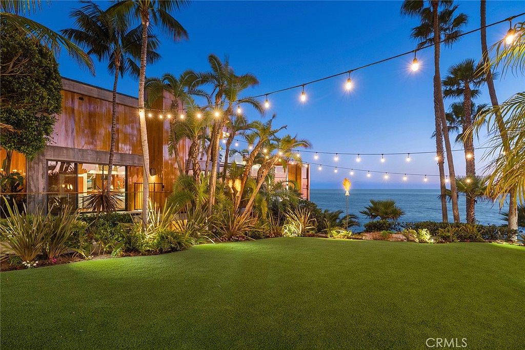 This stunning twilight view captures a lush, manicured lawn overlooking the ocean, framed by mature palm trees and string lights. The modern home features warm wood-toned siding and large glass windows that reflect the evening sky. The perspective is low and wide, emphasizing the seamless transition between the private garden and the expansive coastal horizon.