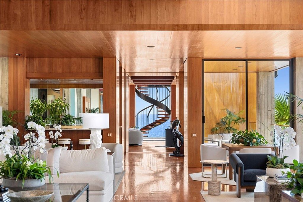 This sophisticated living space features warm wood-paneled walls and ceilings, creating a cohesive and luxurious atmosphere. A striking spiral staircase serves as a focal point, leading the eye toward a stunning ocean view framed by large glass panels. The room is furnished with plush, light-toned seating and accented with elegant floral arrangements, blending modern architectural design with a serene, coastal aesthetic.