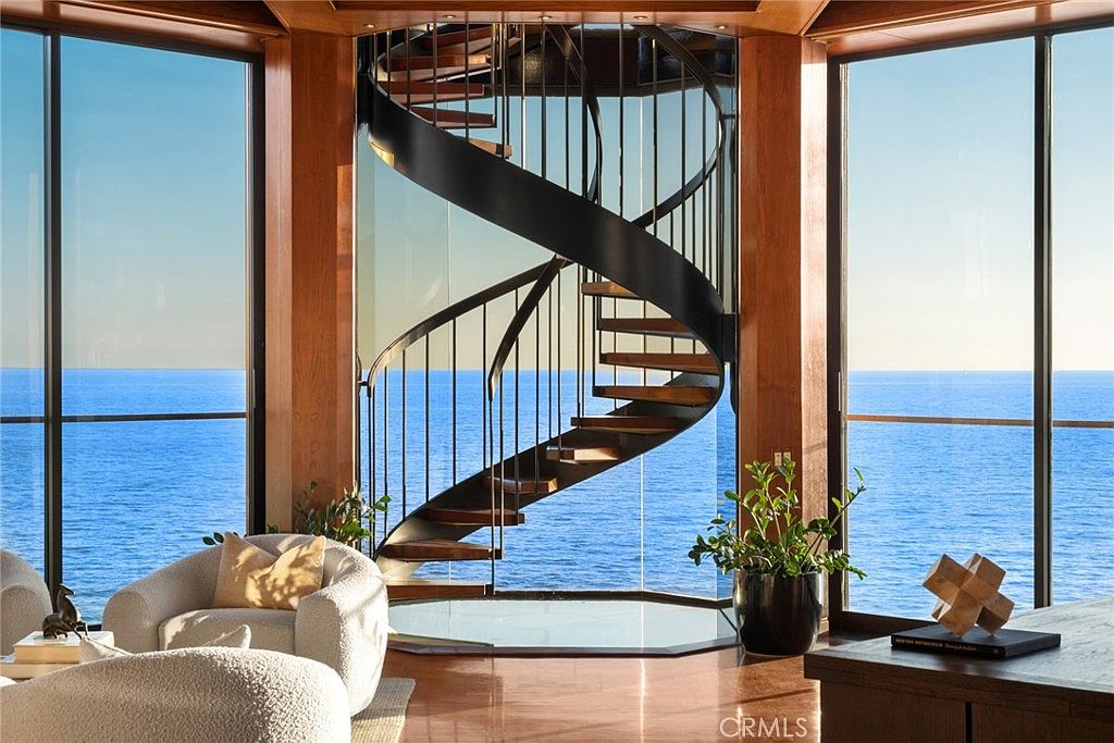 This sophisticated living space features a striking spiral staircase with dark metal railings and wood treads as its central focal point. The room is framed by expansive floor-to-ceiling windows that offer an unobstructed, panoramic view of the ocean. The interior is finished with warm wood flooring, comfortable textured seating, and a potted plant, creating a serene and luxurious atmosphere.