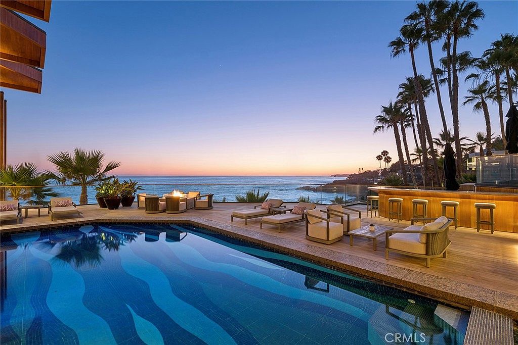 This luxurious outdoor living space features a custom-shaped swimming pool with a striking blue wave-patterned tile design. The expansive deck is furnished with multiple lounge chairs, a fire pit seating area, and a built-in bar overlooking the ocean at sunset. The scene captures a serene, high-end coastal atmosphere with palm trees framing the horizon.
