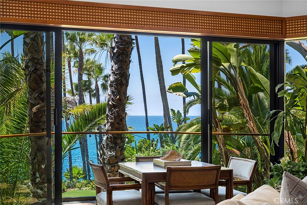 This dining area features a wooden table and chairs positioned in front of large, expansive windows that offer a stunning, unobstructed view of the ocean and lush palm trees. The room is framed by a decorative wooden lattice detail above the glass, creating a warm, tropical atmosphere. The perspective is from inside the home, looking out toward the serene coastal landscape.