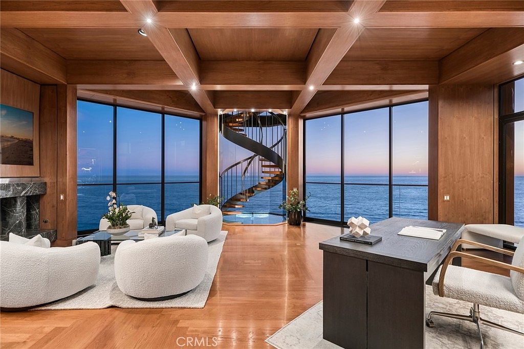 This sophisticated home office features a striking spiral staircase as a central architectural focal point, set against expansive glass walls that offer panoramic ocean views. The room is defined by a coffered wood ceiling and warm wood flooring, creating a luxurious and serene atmosphere. A dark desk sits in the foreground, complemented by plush, rounded seating arranged in a conversational grouping near a stone-clad fireplace.