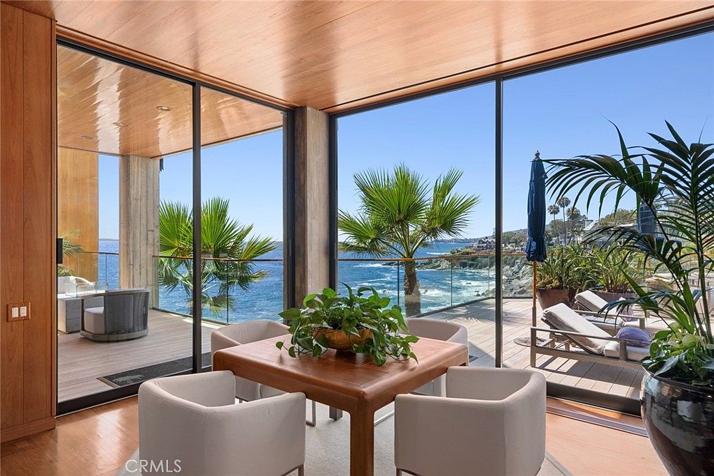 This elegant dining area features a square wooden table surrounded by four modern, light-colored upholstered chairs, centered under a warm wood-paneled ceiling. The space is defined by expansive floor-to-ceiling glass walls that provide an unobstructed, panoramic view of the ocean and rugged coastline. A large potted plant sits on the table, while the adjacent deck area, visible through the glass, features lounge chairs and a blue umbrella, creating a seamless indoor-outdoor living experience.