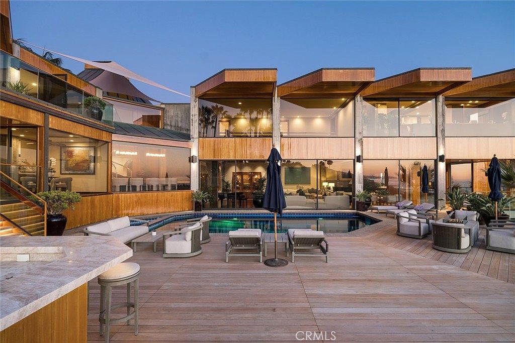 This expansive outdoor living space features a multi-level deck constructed of light wood, centered around a rectangular swimming pool. The home showcases a modern architectural style with large glass walls that provide a seamless transition between the interior living areas and the outdoor deck. The scene is set during the golden hour, creating a warm, inviting atmosphere perfect for entertaining.