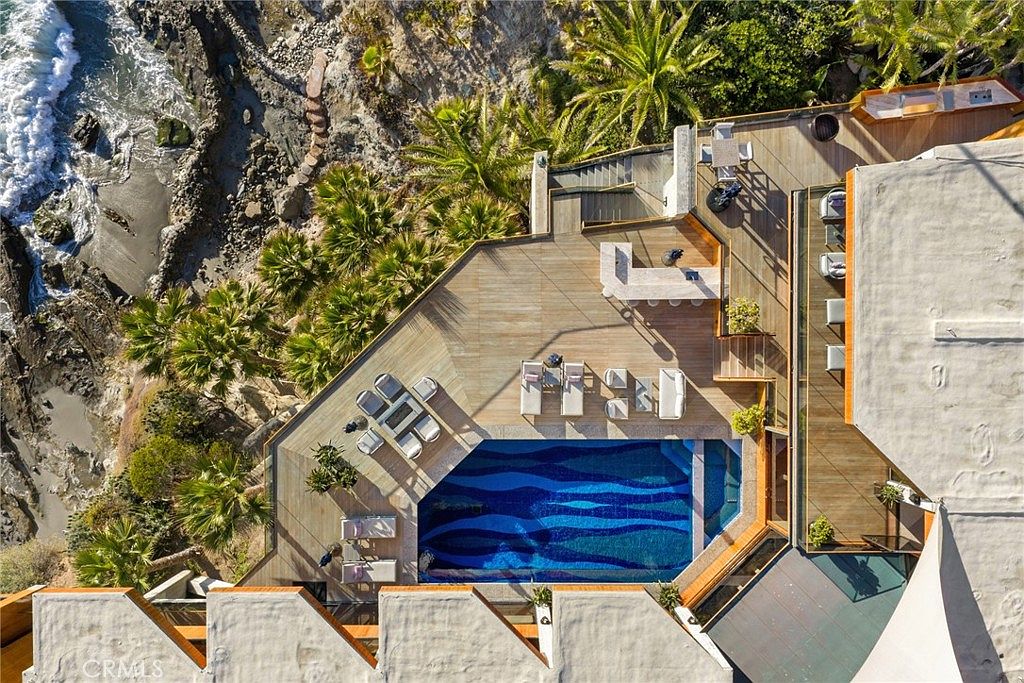This high-angle aerial perspective captures a luxurious coastal property featuring a multi-level wooden deck that integrates a vibrant, mosaic-tiled swimming pool. The outdoor living space is equipped with lounge chairs, a dining area, and a bar, all set against a dramatic backdrop of rugged cliffs and the crashing ocean surf. Lush palm trees and natural landscaping frame the deck, emphasizing the seamless blend of modern architecture and the natural seaside environment.