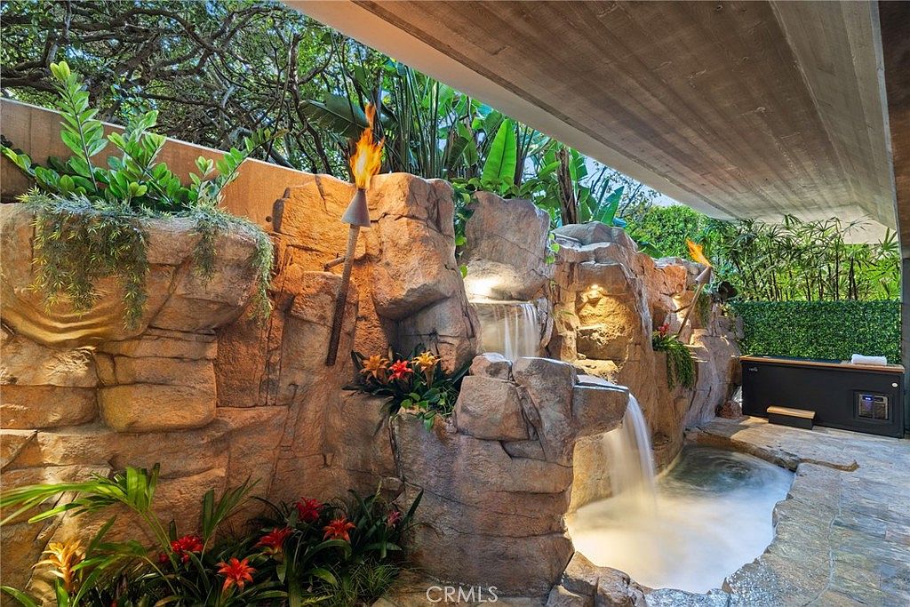 This luxurious outdoor space features a custom-built rock waterfall cascading into a small, tranquil pool. The area is accented by tropical landscaping, tiki-style torches, and a modern hot tub tucked under a concrete overhang. The scene creates a resort-like atmosphere, perfect for relaxation and high-end outdoor living.