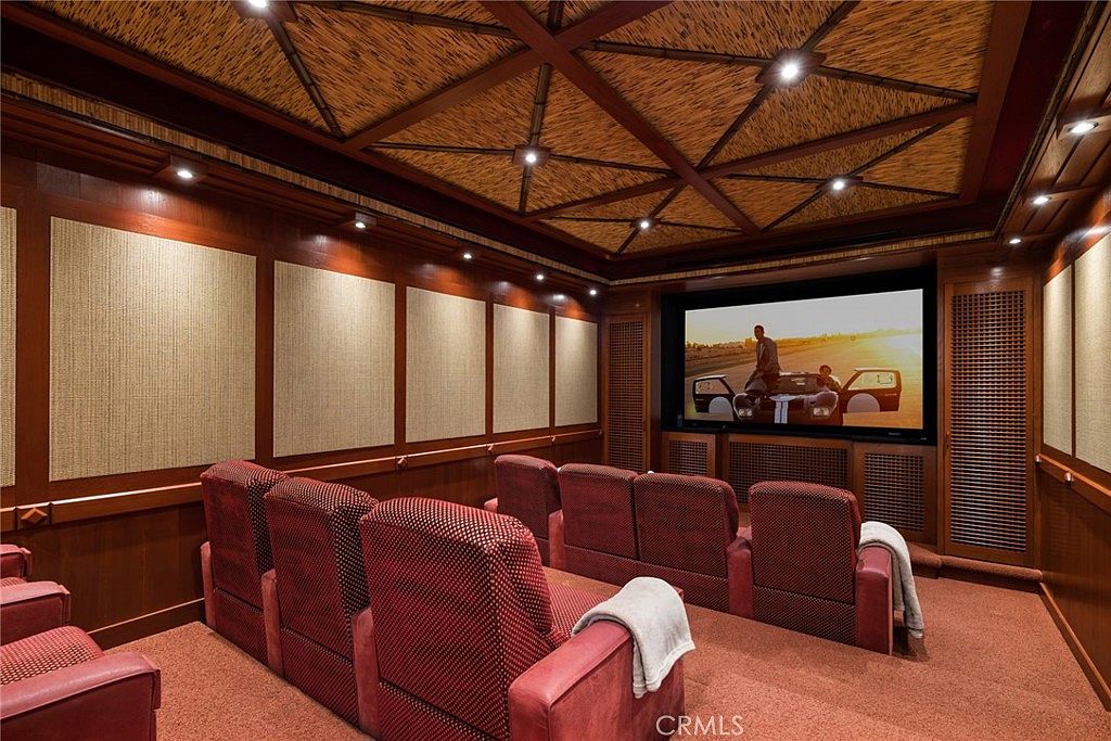 This dedicated home theater room features tiered rows of plush red upholstered seating facing a large integrated screen. The room is characterized by its rich wood-paneled walls and a distinctive textured, woven-style ceiling with recessed lighting. The overall atmosphere is dark, intimate, and designed for an immersive cinematic experience.