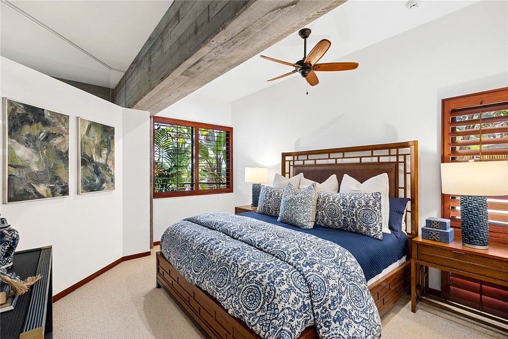 This primary bedroom features a prominent wooden bed frame with a lattice-style headboard, dressed in blue and white patterned bedding. A rustic wooden beam spans the ceiling, adding architectural character to the room, while large windows with wooden shutters allow natural light to filter in. The space is decorated with two framed abstract artworks on the wall and matching bedside tables with lamps, creating a cohesive and tranquil atmosphere.