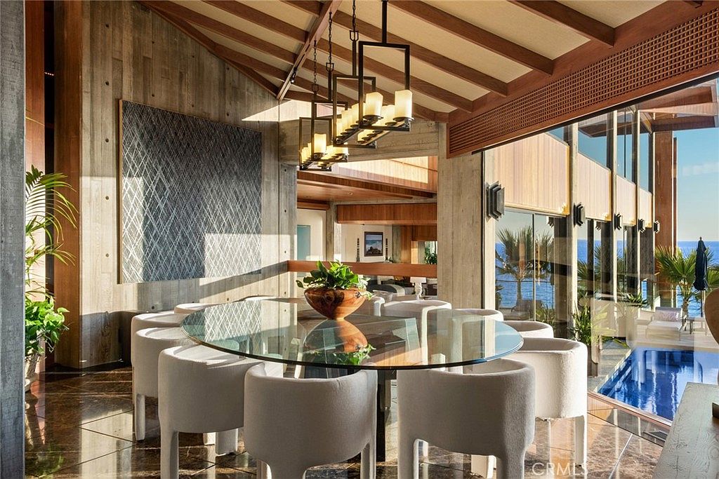 This sophisticated dining area features a large glass-topped table surrounded by modern, cream-colored upholstered chairs, centered beneath a striking multi-tiered rectangular chandelier. The space is defined by warm wood-paneled ceilings and textured concrete walls, creating a blend of industrial and organic aesthetics. Large windows provide an expansive view of the ocean and an adjacent swimming pool, bathing the room in natural light and emphasizing the seamless connection between indoor and outdoor living.